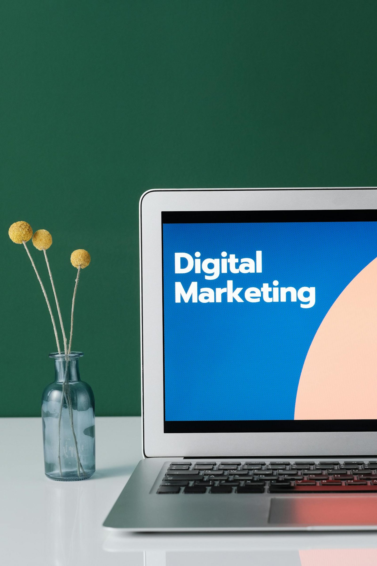 A laptop displaying 'Digital Marketing' with a minimalist floral decor in a glass vase.